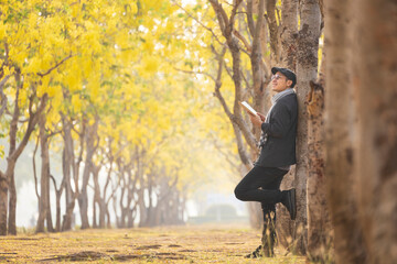 Asian man wearing sweater reading the book while leaning on the yellow leaves ginkgo tree in the...