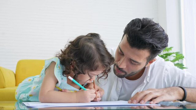 Happy family father teach little daughter using color pencil drawing on paper book in living room. Dad and cute child girl kid enjoy and having fun leisure activity and homeschooling together at home