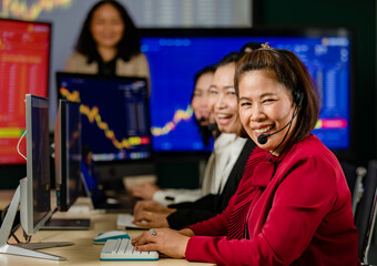 Fototapeta premium Group of Asian female customer service operators wears microphone headsets sit smiling in front daily graph chart computer monitor screen talking and trading buy sell stock market online with client
