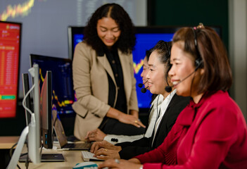 Group of Asian female customer service operators wears microphone headsets sit smiling in front daily graph chart computer monitor screen talking and trading buy sell stock market online with client