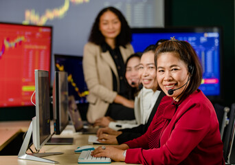 Asian female professional stock exchange manager sit on desk smile in front chart monitor look at camera while customer service operator team wears microphone headsets working in blurred foreground