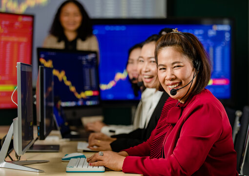 Group of Asian female customer service operators wears microphone headsets sit smiling in front daily graph chart computer monitor screen talking and trading buy sell stock market online with client