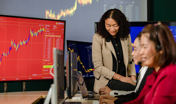 Group of Asian female customer service operators wears microphone headsets sit smiling in front daily graph chart computer monitor screen talking and trading buy sell stock market online with client