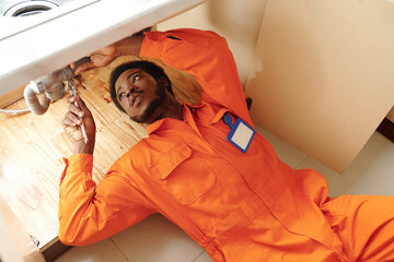 Young plumber in orange uniform lying under sink and fixing leaking pipe