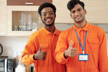 Team of positive plumbers in overalls smiling and showing thumbs-up when standing in kitchen