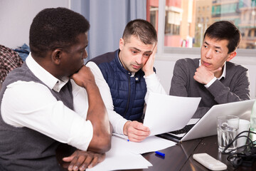 Three upset male friends looking worriedly at papers at home table