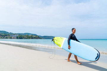 Obraz premium Confidence Asian man in wetsuit holding surfboard and walking on tropical beach at summer sunny day. Healthy sportsman enjoy outdoor active lifestyle play extreme sport surfing on summer vacation