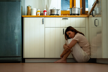 Depressed Asian woman sitting on the floor in kitchen and hugging herself with sadness. Loneliness...