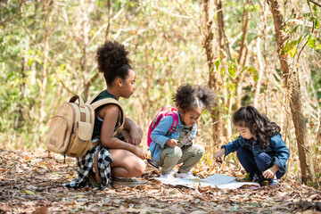 Group of Diversity little girl friends with backpack hiking together at forest mountain in summer sunny day. Three kids having fun outdoor activity sitting and looking at the map exploring the forest.