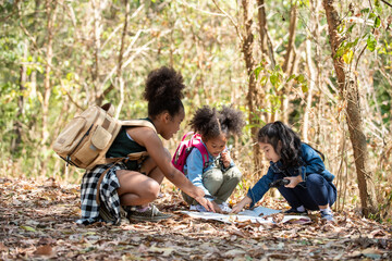 Group of Diversity little girl friends with backpack hiking together at forest mountain in summer sunny day. Three kids having fun outdoor activity sitting and looking at the map exploring the forest. - obrazy, fototapety, plakaty