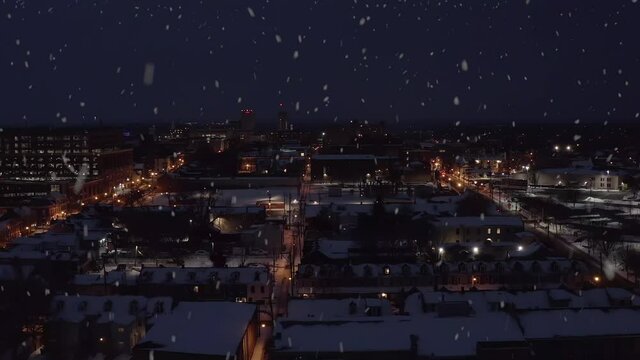 Snow falling at night. Rising aerial of urban city in USA during dark evening. Snow covers rooftops. Snowy cold establishing shot for town in United States.
