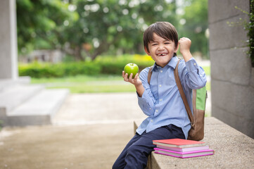 Fototapeta premium A cute little boy wearing a blue shirt and carrying a student backpack holding apples and showing bicep muscles.Child with confidence ready to defense from bullying.Ready to go school again.
