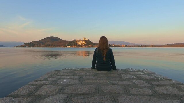 Back view of child girl sitting on pier edge looks at Angera castle on Maggiore lake in Italy at sunset. First-person pov