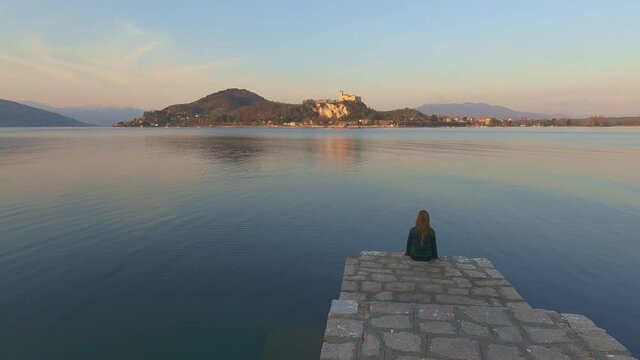 Back view of child girl sitting on pier edge looks at Angera castle overlooking Maggiore lake in Italy at sunset. Slow-motion