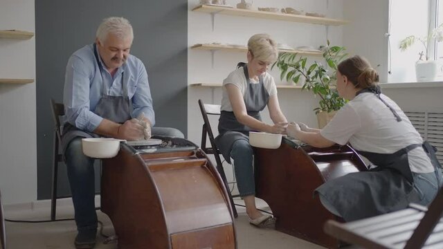 A female potter's wheel sculpting teacher explains how to work and teaches an elderly woman to work with clay and make mugs and jugs. Master class for pensioners. Pottery courses