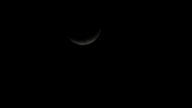 Moon, in its Crescent phase covered with clouds moving down diagonally to the right side of the frame as it is coming in and out in the dark of the night.