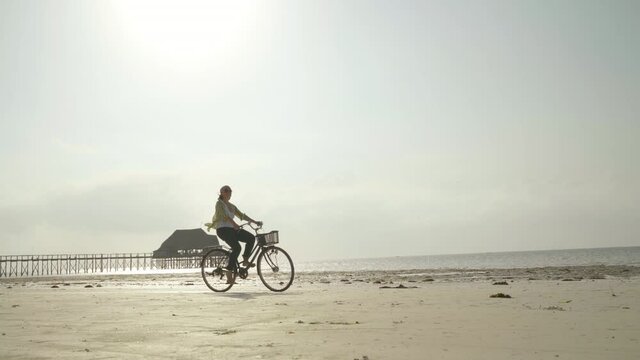 4K static footage woman riding vintage city bicycle on empty sandy beach near sea and pier in background. Resort vacation, beach holiday, tourist concept.