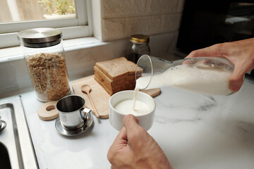 Hands of man pouring milk in cup of coffee when making breakfast in the morning