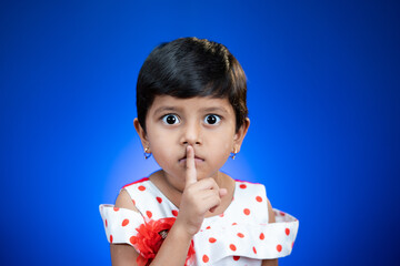 Cute girl kid saying keep silence quite by placing finger on mouth on blue studio background by...