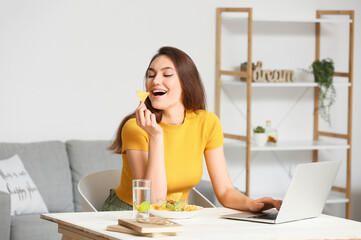 Beautiful young woman with laptop eating tasty nachos at home