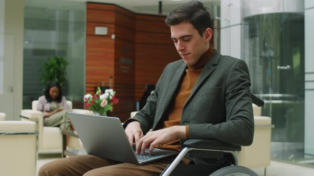 Young disabled businessman sitting in wheelchair and using laptop while working in modern office center