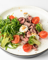 Healthy salad made from meat, cherry tomatoes, broccoli, goat cheese and fresh arugula served in a white plate.