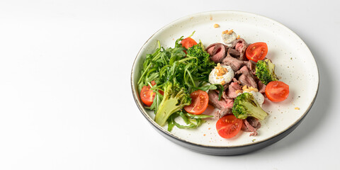 Healthy salad made from meat, cherry tomatoes, broccoli, goat cheese and fresh arugula served in a white plate.