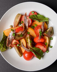 Vegetable salad made of tomatoes and fish slices, served in a white plate over black background.