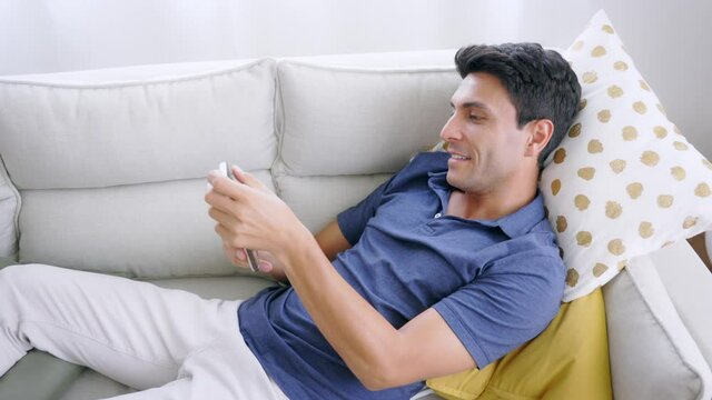 Handsome young caucasian man lying on sofa in a comfortable position playing games and watching video on smartphone during free time at home