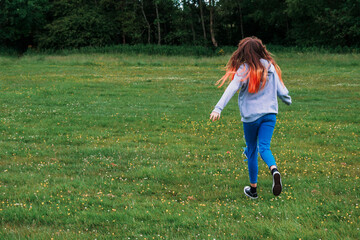 Fototapeta premium Teenager girl with long hair running on a grass in a park. Back to camera. Outdoor activity. The model is tall with long colored hair.