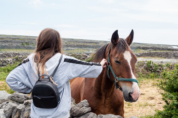 Teenager girl with back pack and brown horse behind dry stone fence. Inishmore, Aran Islands,...