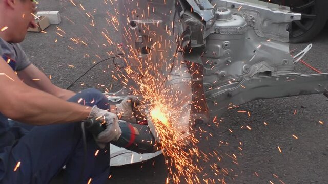 A locksmith cuts out a metal part of a car body with a tool. Dismantling a transport for auto parts in the garage. During operation, many bright yellow sparks fly to the sides.
