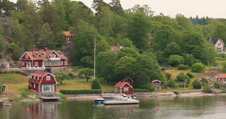 Many Red Swedish Wooden Sauna Logs Cabins Houses On Island Coast In Summer Cloudy Day