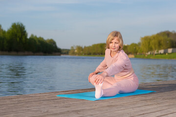 Fototapeta na wymiar Sport Ideas. Mature Caucasian Woman During Yoga Legs Stretching Training On Wooden Stage Near Water Outdoor.
