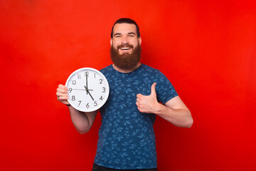 Pleased bearded man holding clock while showing thumb up and looking at the camera over red...