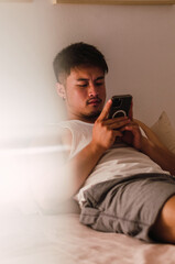 Fototapeta premium Young handsome Chinese man at home lying in bed using smartphone