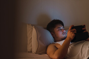 Fototapeta premium Young handsome Chinese man at home lying in bed using digital tablet