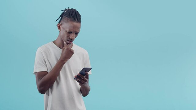 Black puzzled man rubs chin with hand, using phone, and indignantly talking in the studio on a blue background.