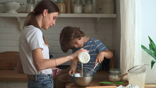 Young mom and son making dough and standing in home kitchen indoors.