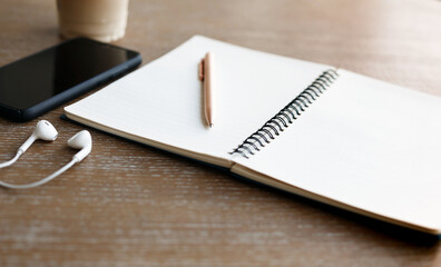 Focus at pen on blank notebook on wooden table with mobile, headphone and coffee
