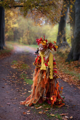 Herbstfee, ein kleines blondes Mädchen mit dem bunten Herbstkranz auf dem Kopf steht im bunten Herbstwald. Das Gesicht ist mit der Herbstmuster geschminkt.. In der Hand ist eine Kastanie.
