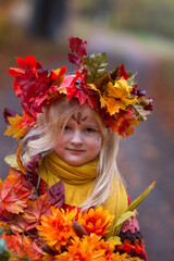 Herbstfee, ein kleines blondes Mädchen mit dem bunten Herbstkranz auf dem Kopf steht im bunten Herbstwald. Das Gesicht ist mit der Herbstmuster geschminkt. Sie hält seht viele bunte Blätter 
