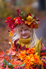 Herbstfee, ein kleines blondes Mädchen mit dem bunten Herbstkranz auf dem Kopf steht im bunten Herbstwald. Das Gesicht ist mit der Herbstmuster geschminkt. Sie hält seht viele bunte Blätter 