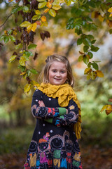 Süßes, kleines, blondes Mädchen steht im Wald. Sie hat ihre Hände überkreuzt. Sie lächelt. Im Vordergrund ist der Herbstbaum. Im Hintergrund sind verschwommene Bäume.