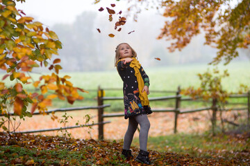 Blondes kleines Mädchen spielt im Wald mit den Herbstblättern.Sie schmeißt die Blätter hoch. Hält ihre Hand ausgestreckt und fängt die bunte Herbstblätter.Im Hintergrund ist ein Feld, Holzzaun, Nebel.