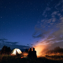 Fototapeta na wymiar Beautiful view of night starry sky over grassy hill with illuminated camp tent, campfire and hikers. Back view of couple tourists having a rest near bonfire. Concept of night camping, relationship.