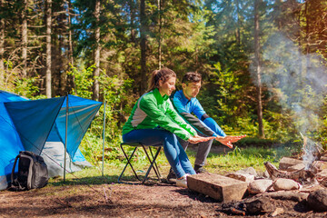 Naklejka na ściany i meble Young couple of travelers enjoy campfire in summer forest. Tourists sitting by tent warming hands. Camping in woods