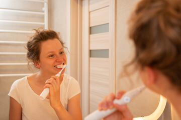 A girl bruhes teeth with white elctric toothbrush in bathroom, looking in the mirror