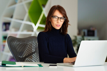 Attractive businesswoman wearing eyeglasses and turtleneck sweater, using laptop while working at...