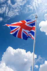 Union Jack Flag. Close-up of a national UK flag with flagpole, blowing in the wind on a blue sky with clouds and copy space.
